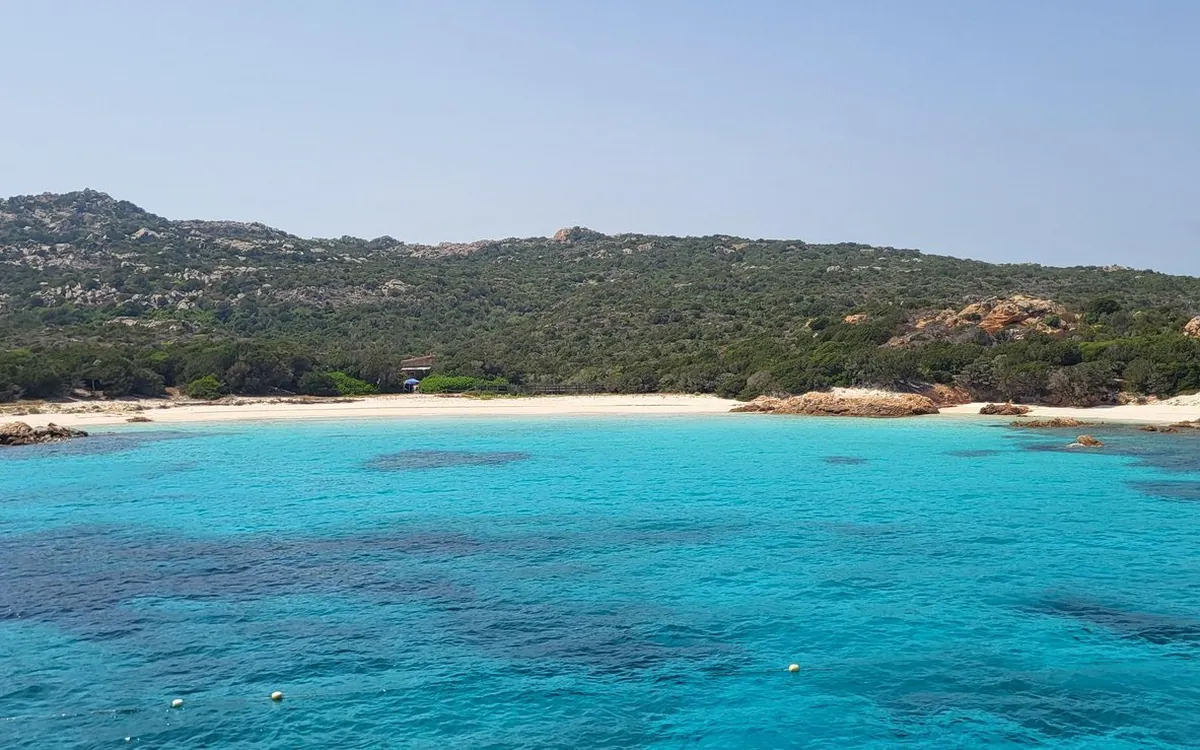 A long white beach below a green hillside, seen from turquoise water