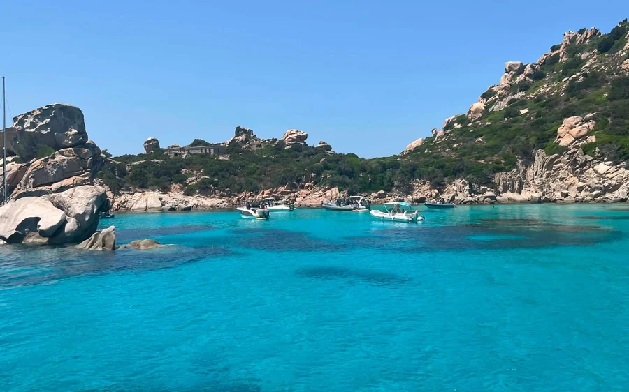 Boats anchored in a turquoise cove between granite rocks and a rocky hill