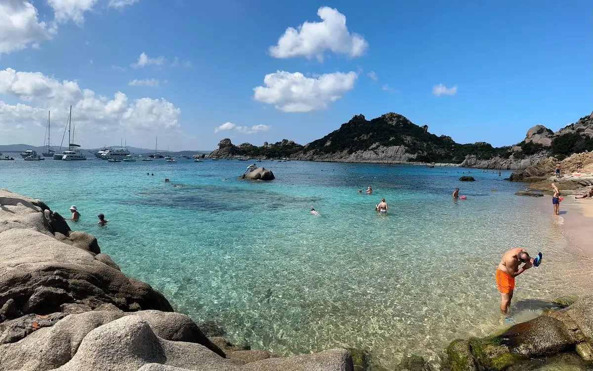 A man in orange shorts bending over a mask in knee-deep clear water at the edge of a granite cove