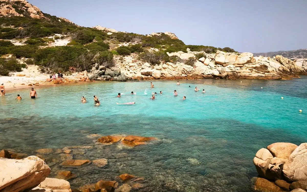 Swimmers in a turquoise cove with orange rocks in front and pale boulders on the shore
