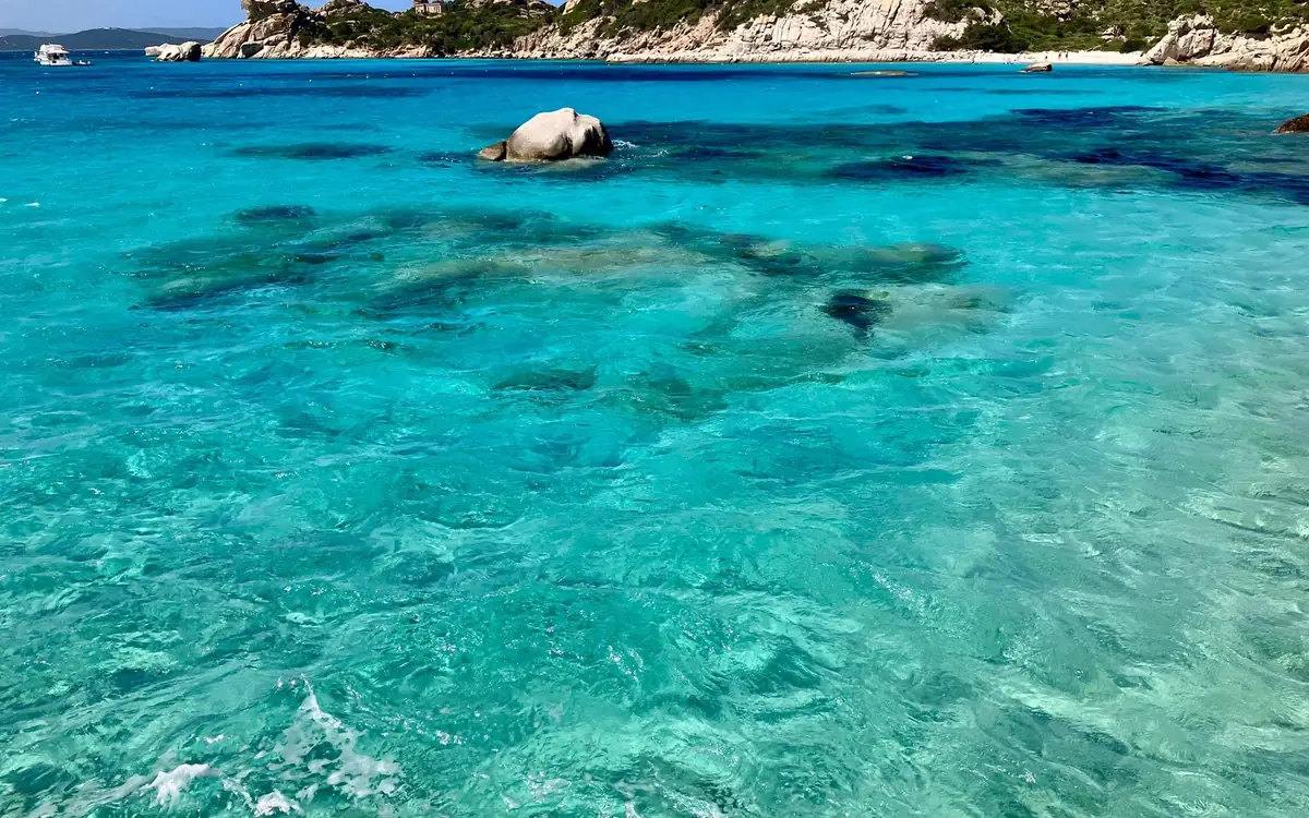 Bright turquoise shallows with dark patches of seagrass and a round granite rock, white cliffs behind