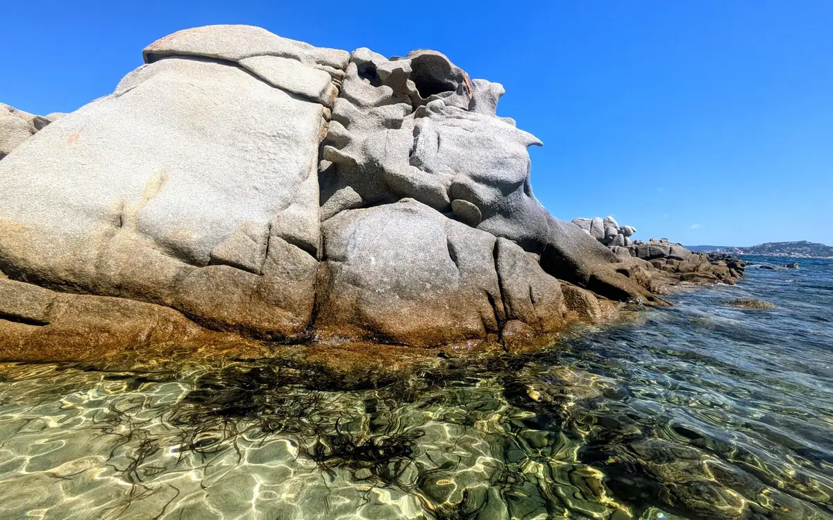 A grey wind-worn granite boulder above clear shallows with dark seaweed on the sand
