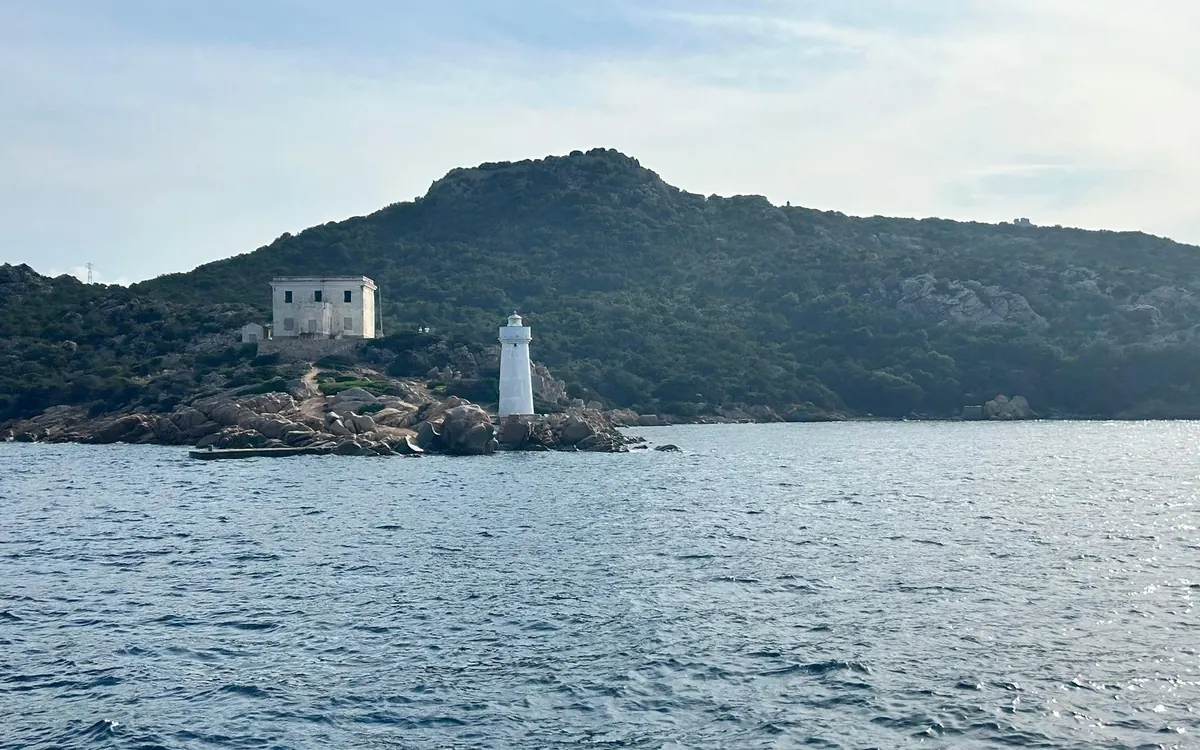 A white lighthouse and a square white building on a rocky point under a wooded hill